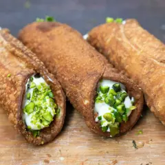 Trzy chrupiące włoskie desery cannoli, wypełnione kremem i posypane pistacjami, kuszą na drewnianej desce.