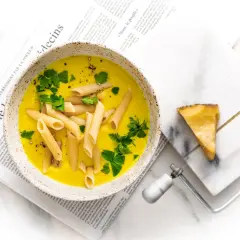 Zupa neapolitańska z makaronem penne i natką pietruszki, podana na gazecie. Obok kawałek sera z ostrzem do sera.