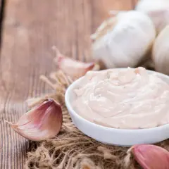 Kremowy sos aioli w miseczce, otoczony świeżymi ząbkami czosnku i całymi główkami na drewnianym tle.
