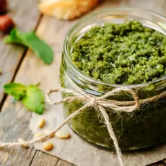 Słoik domowego pesto z bazylii, ozdobiony sznurkiem. Obok leżą orzeszki piniowe i listki bazylii.