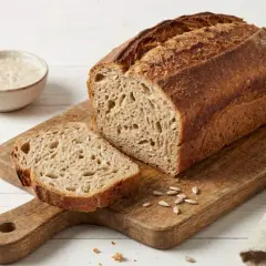 Pyszny, świeży chleb orkiszowy z chrupiącą skórką, pokrojony na desce, obok miseczki z zakwasem i ziarnami.