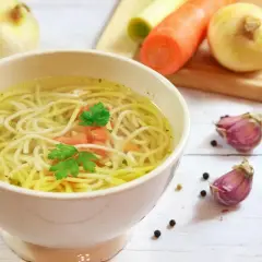 Misternie ułożone nitki makaronu w rosole, z dodatkiem marchewki i natki. Obok cebula, czosnek i makaron – idealne składniki, by wiedzieć, kiedy dodać warzywa do rosołu.