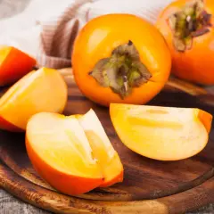 Pomarańczowe owoce kaki, całe i pokrojone w ćwiartki, na drewnianej desce. Odkryj owoc kaki właściwości zdrowotne.