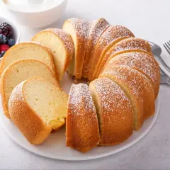 Babka piaskowa przepis babci, puszysta, pokrojona, posypana cukrem pudrem, z owocami i śmietaną w tle.