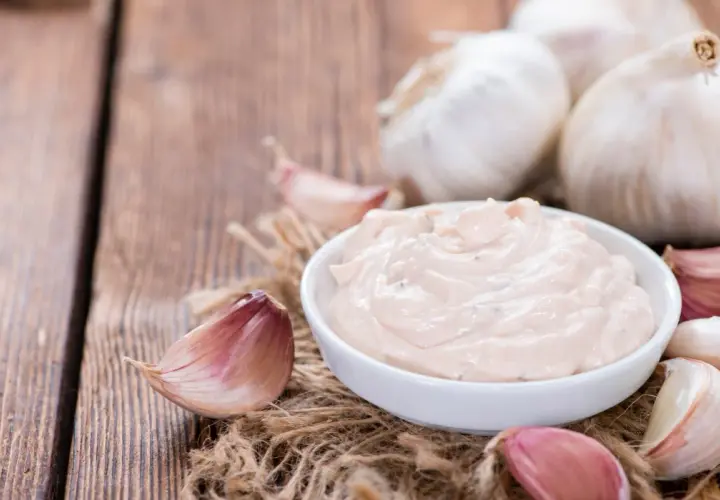 Kremowy sos aioli w miseczce, otoczony świeżymi ząbkami czosnku i całymi główkami na drewnianym tle.
