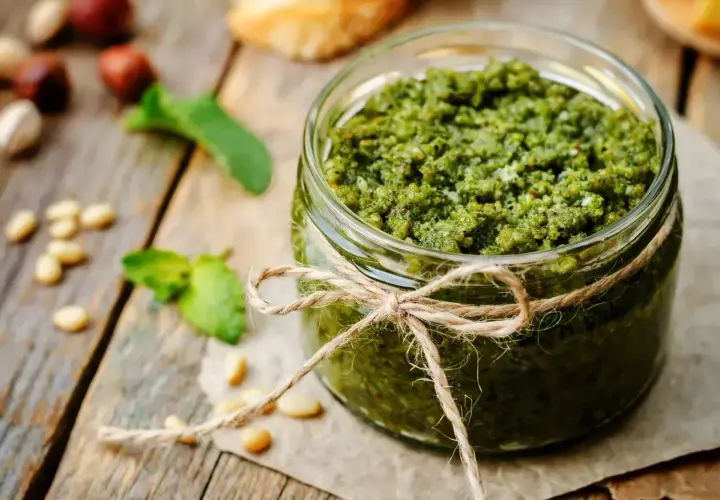Słoik domowego pesto z bazylii, ozdobiony sznurkiem. Obok leżą orzeszki piniowe i listki bazylii.