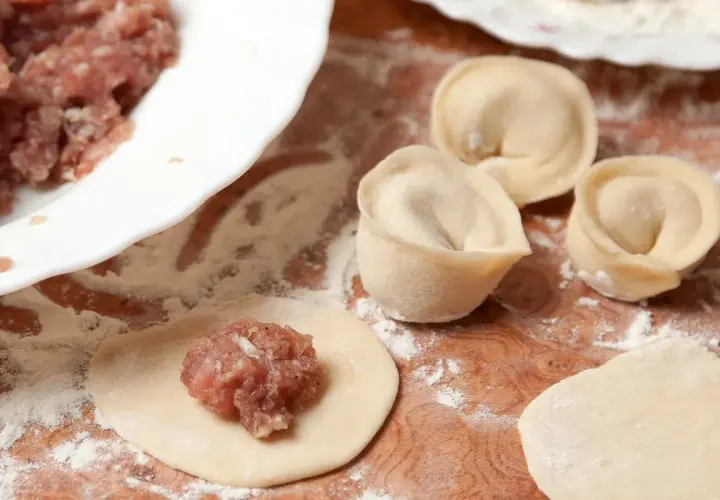 Przygotowanie pierogów z mięsem mielonym surowym. Na drewnianej desce leżą surowe pierogi, obok miska z farszem.