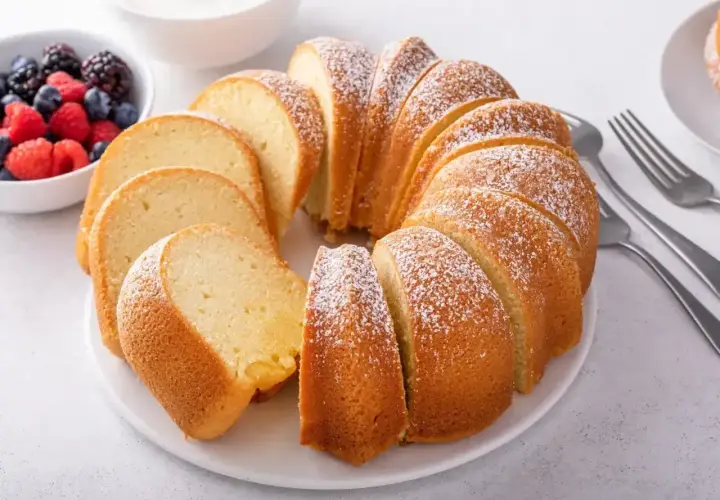 Babka piaskowa przepis babci, puszysta, pokrojona, posypana cukrem pudrem, z owocami i śmietaną w tle.