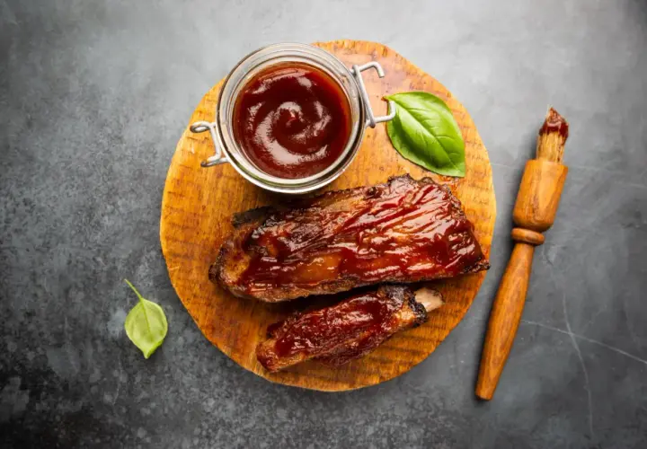 Soczyste żeberka w sosie barbecue, gotowe do podania. Obok słoiczek z sosem do steka i pędzelek.