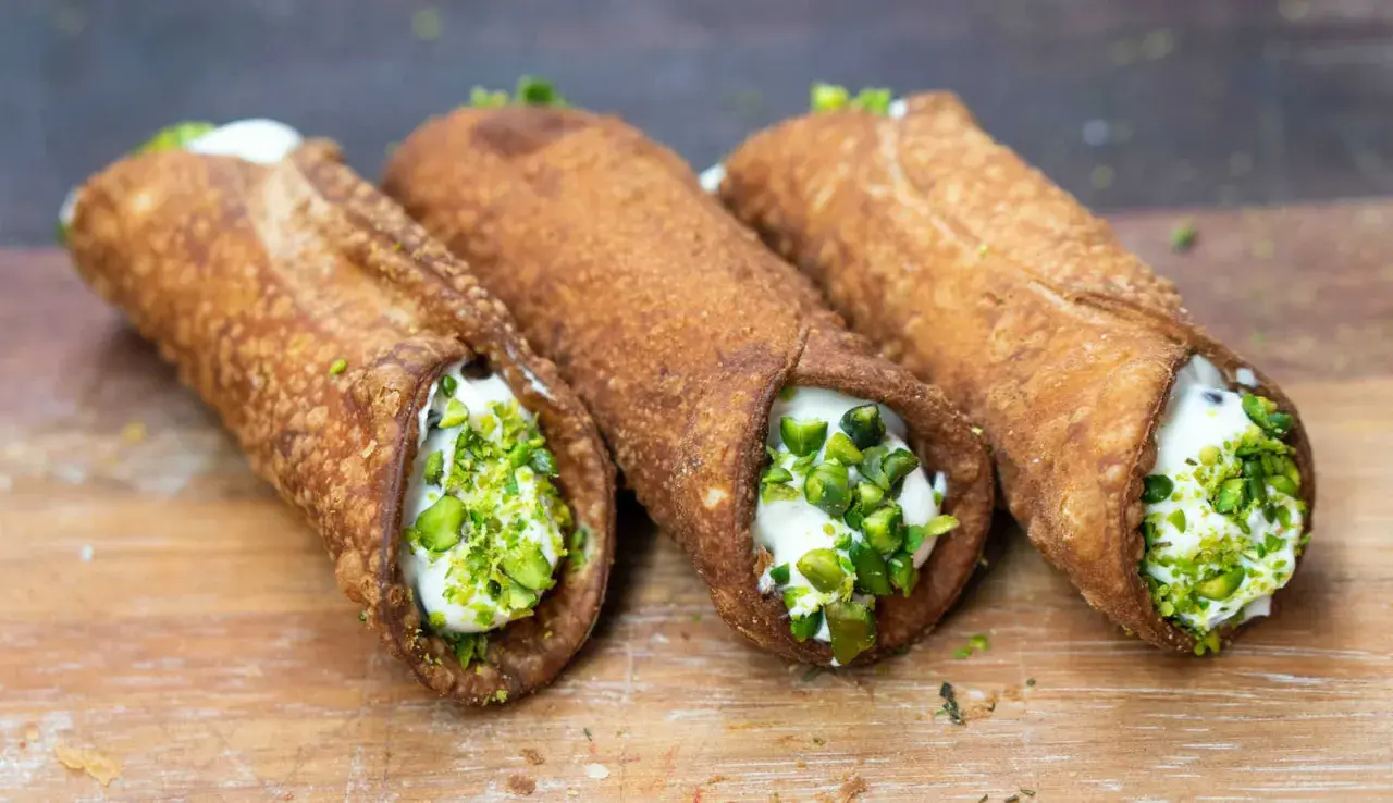 Trzy chrupiące włoskie desery cannoli, wypełnione kremem i posypane pistacjami, kuszą na drewnianej desce.