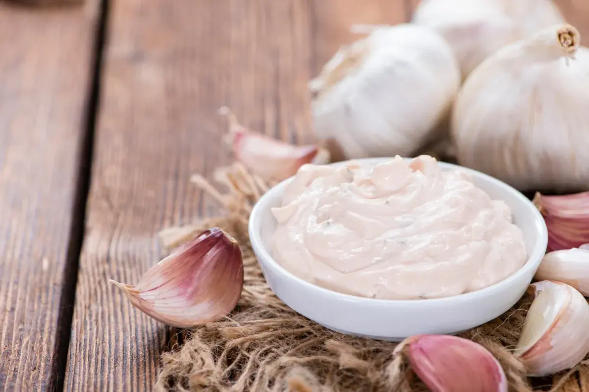 Kremowy sos aioli w miseczce, otoczony świeżymi ząbkami czosnku i całymi główkami na drewnianym tle.