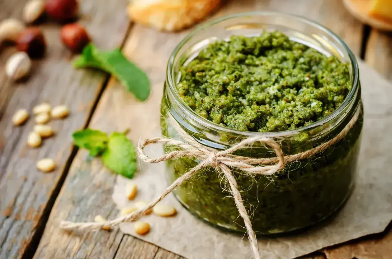 Słoik domowego pesto z bazylii, ozdobiony sznurkiem. Obok leżą orzeszki piniowe i listki bazylii.