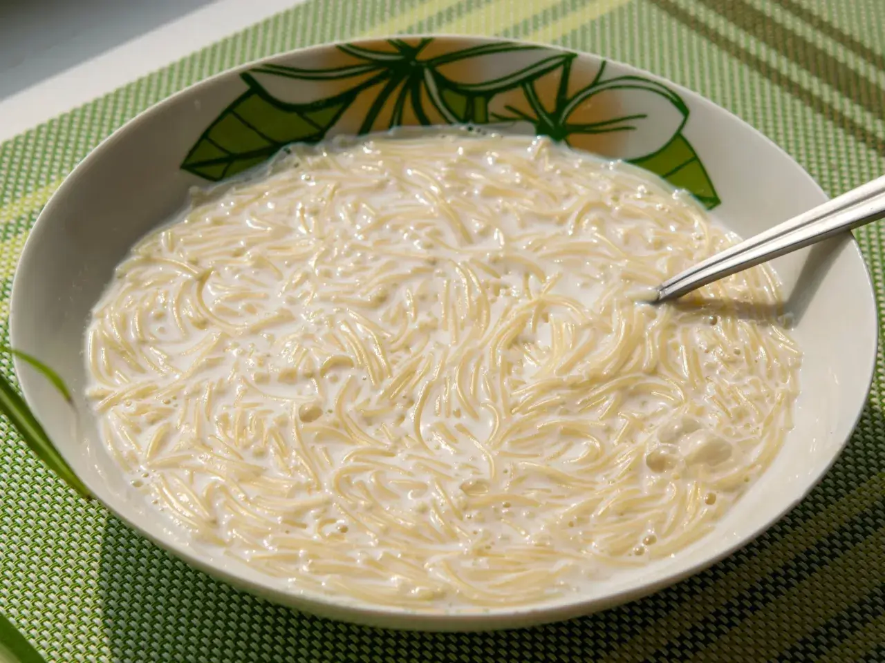 Zupa mleczna z makaronem, podana w misce z motywem kwiatowym. Widoczna łyżeczka zanurzona w kremowym daniu.