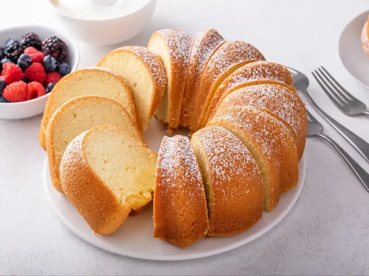 Babka piaskowa przepis babci, puszysta, pokrojona, posypana cukrem pudrem, z owocami i śmietaną w tle.