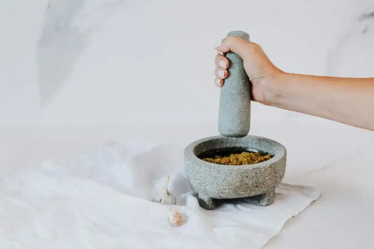 Dłoń uciera w moździerzu aromatyczne pesto z bazylii, obok leżą ząbki czosnku.