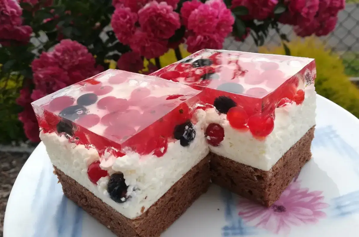Dwa kawałki biszkoptu z galaretką i kremem mascarpone, ozdobione owocami leśnymi, na tle kwitnących róż.