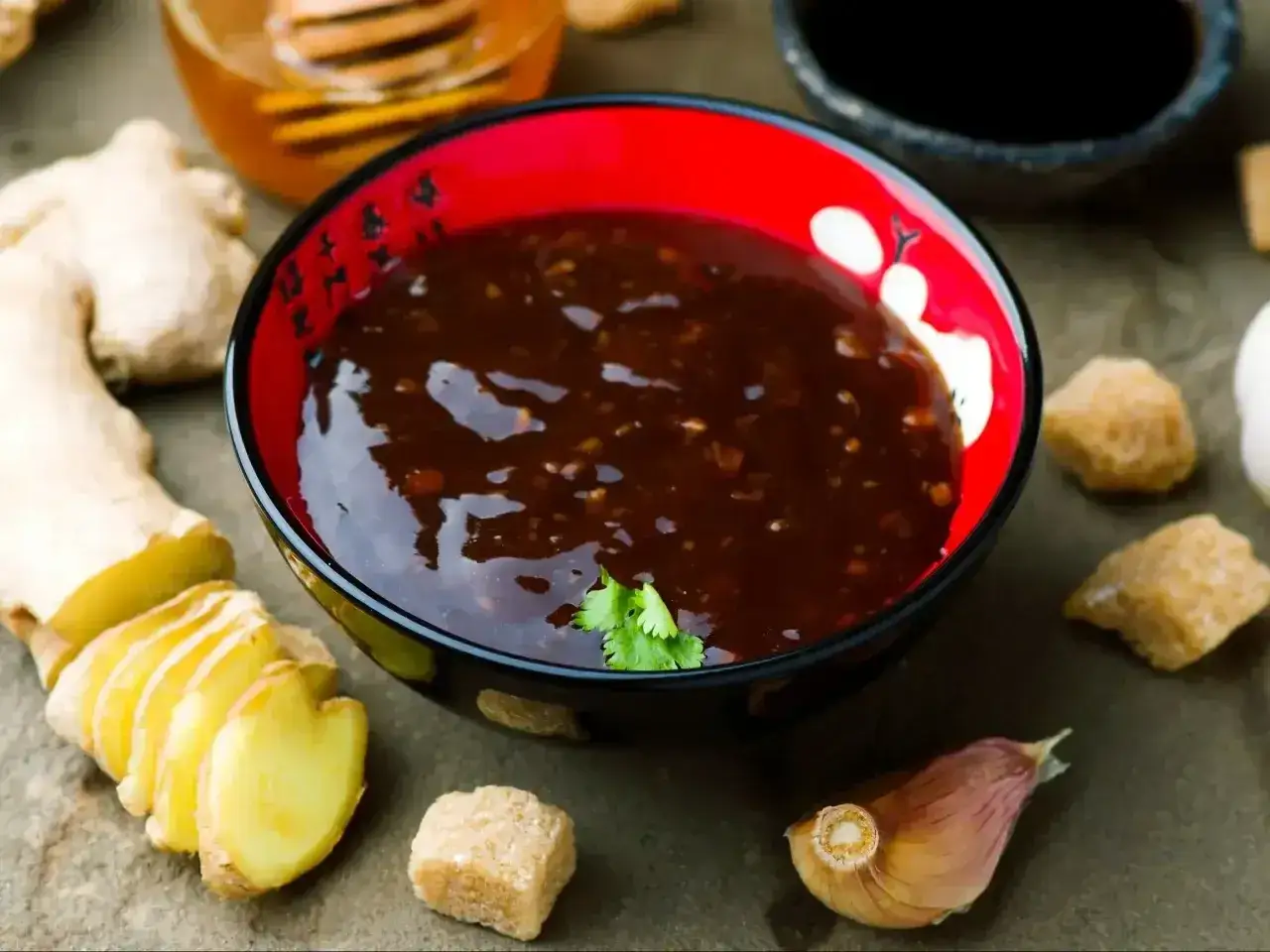 Sos teriyaki przepis: miseczka z ciemnym sosem, obok imbir, czosnek, brązowy cukier i miód. Idealne składniki do domowego sosu teriyaki.