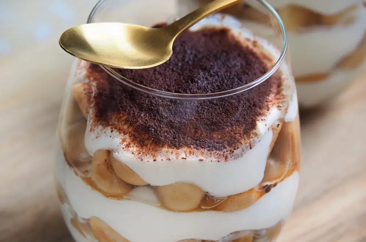 Pyszne tiramisu z mascarpone w pucharku, posypane kakao. Złota łyżeczka czeka na pierwszy kęs tego sprawdzonego przepisu.
