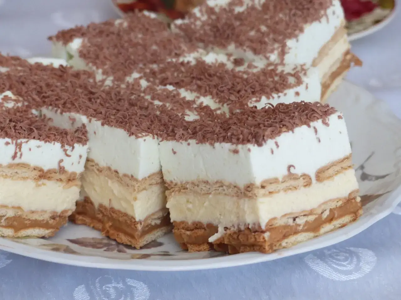 Szybkie ciasto bez pieczenia z budyniem i herbatnikami, przełożone kremem karmelowym, posypane wiórkami czekolady.