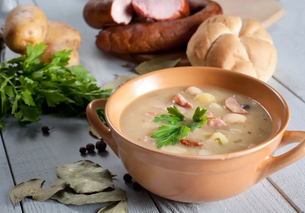Aksamitna zupa fasolowa mojej mamy, z kawałkami kiełbasy i ziemniaków, udekorowana natką pietruszki. Obok świeże ziemniaki, bułka i wędlina.